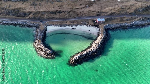 Aerial view of a heart shaped breakwater bay with turquoise water, a red cabin and a scenic coastal road, Lofoten islands Norway, 4K 60fs horizontal Fly Up and Tilt Down drone shot in a sunny day