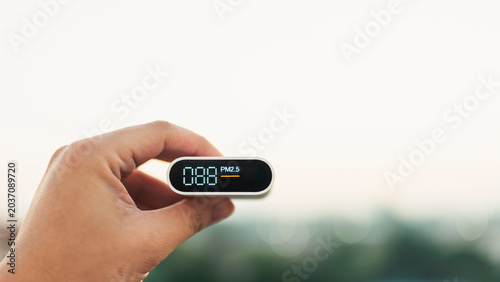 Hand holding a portable air quality monitor showing PM2.5 levels against a blurred outdoor background. Concept of environmental pollution, health care, and smog measurement device.