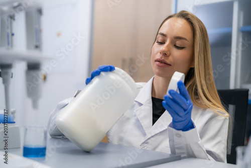 A technician examines a white container of powder while holding a lid at a pharmaceutical workstation. The workspace includes various lab equipment and a blue liquid sample in the background.