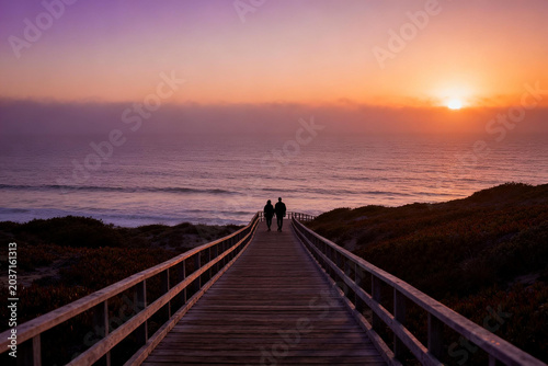 Couple Walking on Wooden Boardwalk Towards Ocean at Sunset