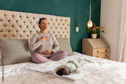 Mid 30s mother and infant daughter sitting cross-legged meditating on bed with tufted headboard