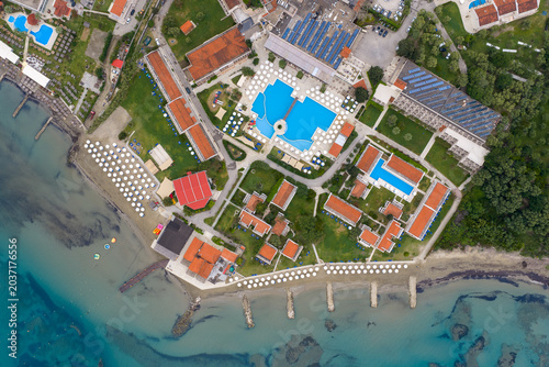 Top down aerial view of a luxury summer resort at Roda Beach in Corfu, Greece, featuring large swimming pools, orange roof buildings, sandy beach with sunbeds, and crystal clear turquoise sea.