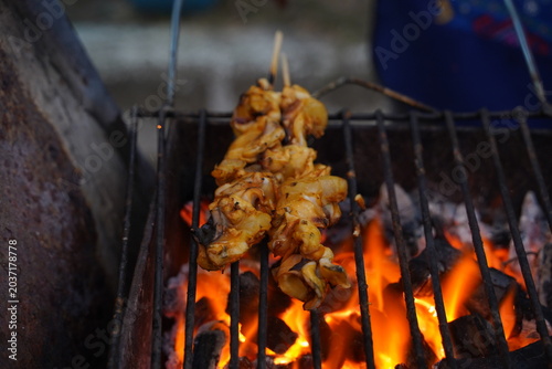 Close up of a yellow marinated squid on a bamboo stick being grilled over a hot flame and glowing charcoal at a street food market.