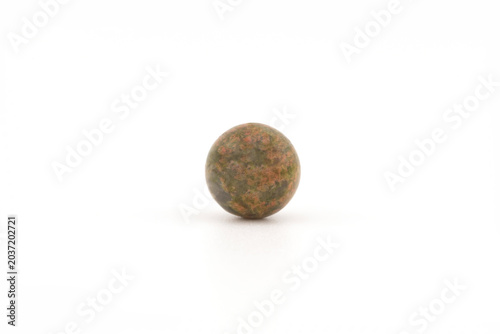 A round unakite stone rests on a white surface. The stone shows green and pink colors. Light shines on it, highlighting its smooth surface and natural patterns.