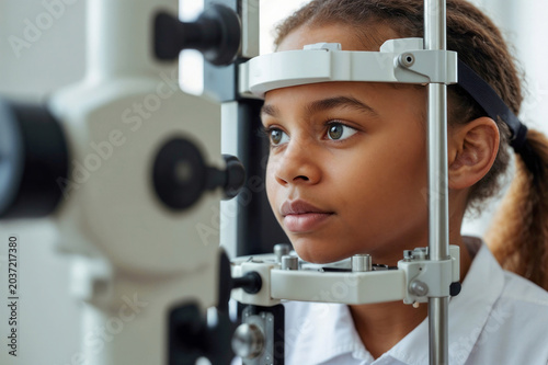 Portrait of black Child gets eye exam with special machine. Young black girl looks into ophthalmic device at clinic