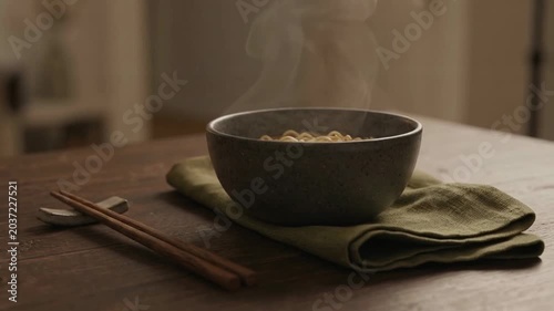 A steaming bowl of noodles placed on a wooden table with chopsticks and a green napkin, creating a cozy atmosphere