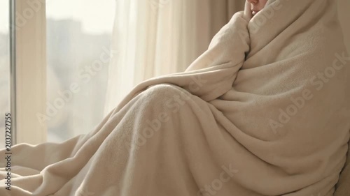 A person wrapped in a cozy beige blanket sitting by a window, evoking feelings of comfort and introspection
