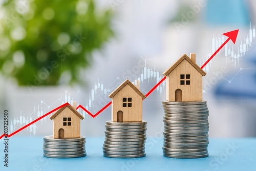 Miniature wooden houses are stacked on coins with a rising graph in the background, illustrating real estate investment growth and value.