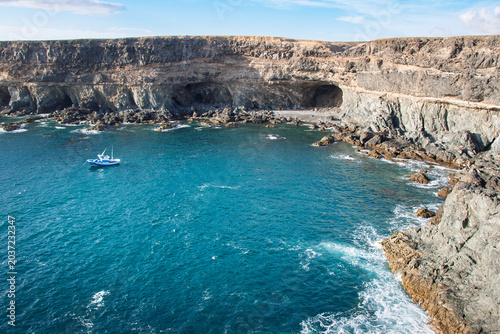 Small boat navigating the turquoise waters of the Atlantic Ocean near rugged volcanic cliffs and hidden sea caves along the picturesque coastline of Fuerteventura, Canary Islands