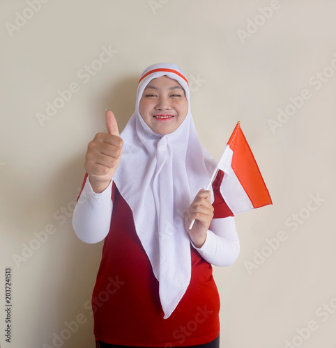 Beautiful Asian Muslim Woman Wearing Hijab And Headband, Holding Indonesia Flag and Showing Thumbs Up. Selective Focus, Focus On Face