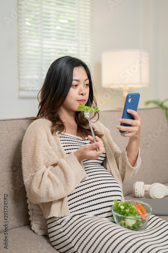 Pregnant asian woman eating vegetables salad looking at phone sitting on sofa in living room at home