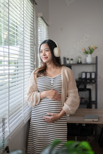 Fotografie Pregnant asian woman listening to music touching belly standing looking out of w