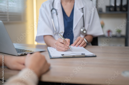 Close up of woman doctor writing in clipboard talking to patient at wooden table in hospital clinic.