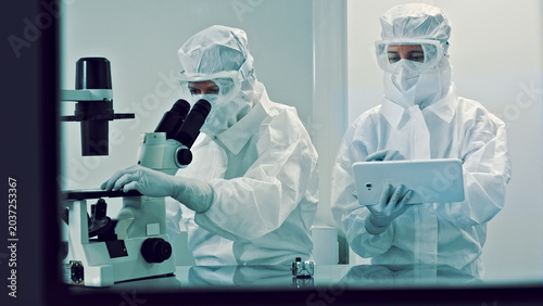 Bild auf Leinwand In a pristine laboratory, two researchers dressed in clean suits meticulously examine a specimen under a microscope, while their colleague analyzes data on a tablet