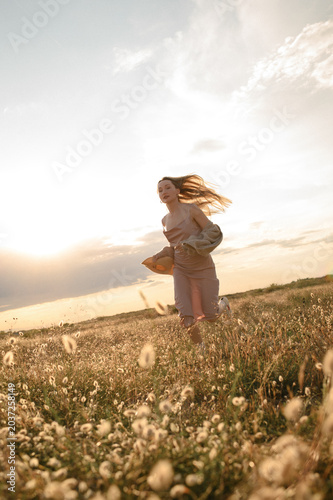 Young woman in dress running through summer field at warm sunset light holding straw hat, long blonde hair flying in wind, freedom, self love and joyful aesthetic lifestyle