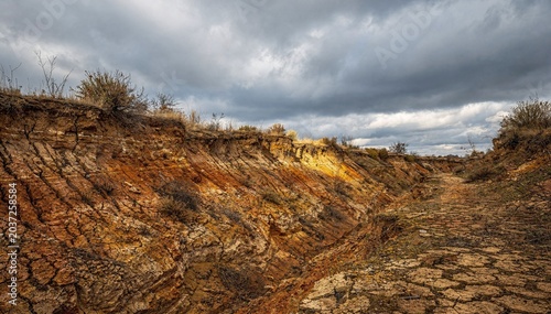 Dramatic landscape of arid brown and orange dirt earth with cracked patterns, eroded slopes, and sparse dry vegetation under a foreboding stormy sky, showcasing a desolate outdoor environment