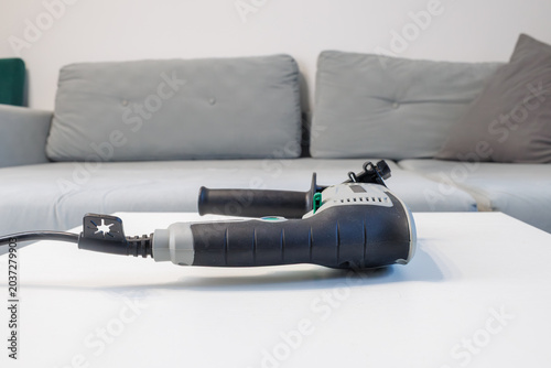 An electric drill lies horizontally on a white table against a blurry gray sofa background