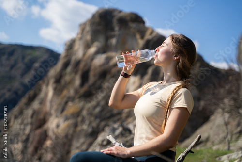 A young sporty woman is hydrating outdoors in a mountainous landscape, sitting and quenching her thirst.
