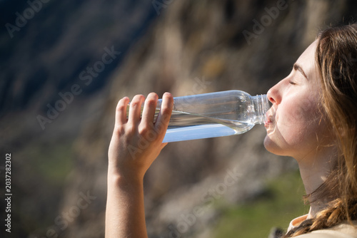 A young woman with closed eyes enjoying drinking water in the sunny mountainous nature, close-up side view portrait.

