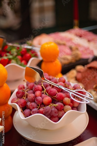 Bowl of red grapes with serving tongs, surrounded by stacked oranges, strawberries, cheese, and charcuterie on a banquet table.