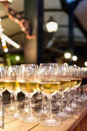 Rows of filled white wine glasses lined up on a wooden bar counter, with warm bokeh string lights glowing in the background at an evening reception.