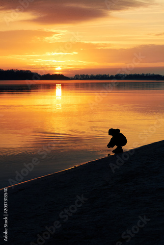 Child silhouette playing by lakeshore during glowing sunset over calm water