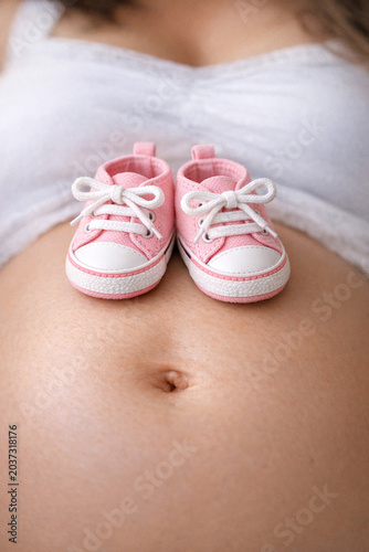 A pregnant woman holds a pair of cute pink baby sneakers with white soles and laces.