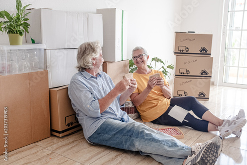 Canvastavla An elderly couple moving into a new home, sitting on the floor for a break and a toast surrounded by moving boxes in an empty room