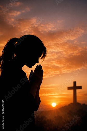 Person in silhouette praying with hands clasped near cross on hilltop during sunset.
