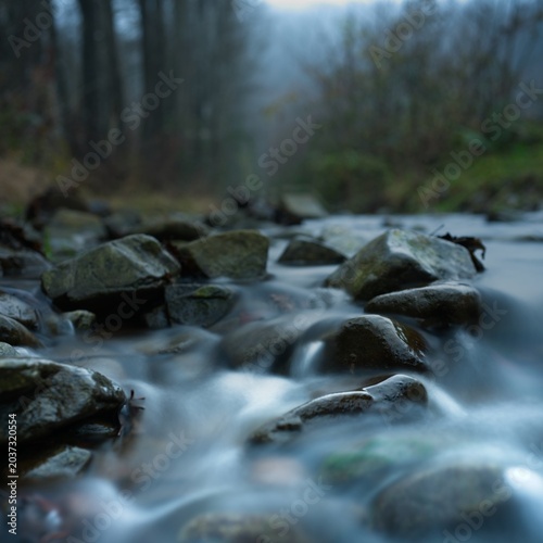 Shallow stream flow between and over stones close up, forest and hazy atmosphere are out of focus in background, calm detail from mountain creek