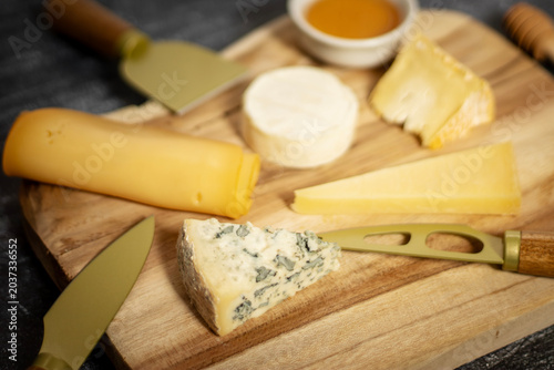Overhead close-up of rustic cheese board with assorted cheeses, honey bowl, and bronze cheese knife on wooden surface, soft light.