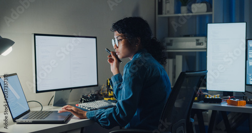 Young African American female programmer coding AI software on multiple screens in night office. Network security, cybersecurity, machine learning, working late, overtime, robotics automation concept.