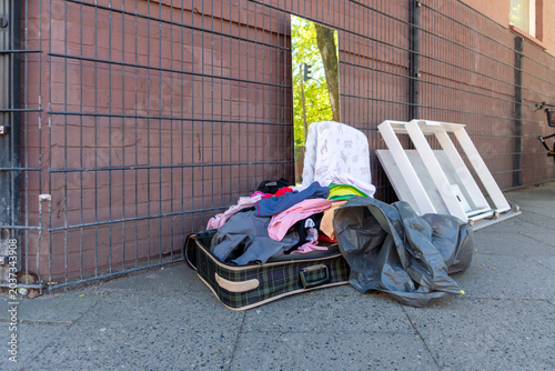 Quadro em tela An open plaid suitcase filled with mixed clothing lies on a city sidewalk beside trash bags and removed window frames, suggesting moving out, eviction, donation, waste, or urban cleanup