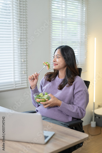 Pretty asian pregnant woman holding bowl eating salad across laptop on table in living room at home.