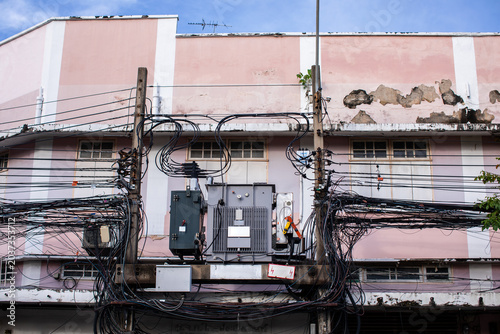 A large electric transformer mounted on concrete poles, tangled in a messy web of overhead power and telecom cables against a weathered pink building facade.