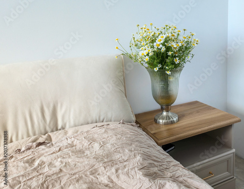 a wide bedroom view showing a glass vase with a fresh chamomile bouquet on a wooden nightstand beside a bed with soft crumpled linen bedding