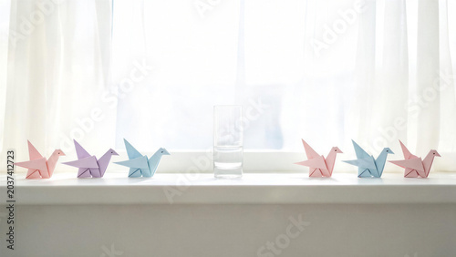 A minimalist arrangement of delicate pastel-colored origami cranes flanking a simple glass of water on a clean white windowsill. An evocative image of purity and peace for the International Day of Pea