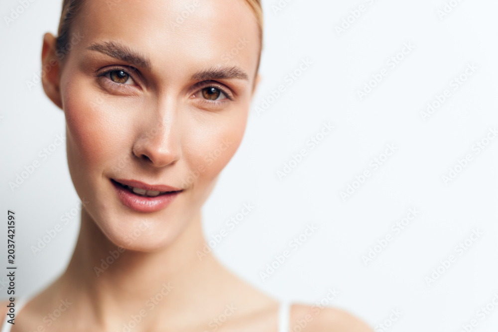 Fototapeta premium Young woman with clear skin and light makeup smiles gently in a close-up portrait against a white background conveying natural beauty and confidence.