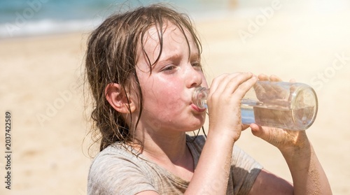  Konzept für extreme Hitze und Dehydration: Ein erschöpftes, stark schwitzendes Mädchen ohne Kopfbedeckung trinkt an einem heißen Strand Wasser, um einem Sonnenstich vorzubeugen.