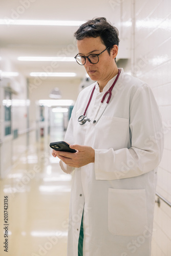 Female doctor using smartphone for patient communication in hospital hallway, modern healthcare technology