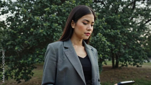 Asian woman in gray blazer walks through a park, glancing at her smartphone while surrounded by lush green trees and natural scenery