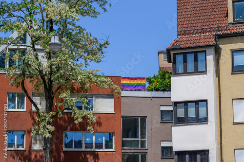 Rainbow flag displayed on rooftop between blooming tree and apartment buildings