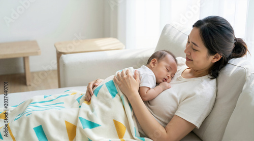 Mother holding her sleeping baby on her chest while sitting on a sofa. Woman embracing her infant covered with a patterned blanket in a bright living room