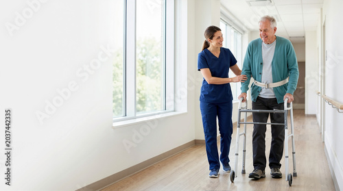 Physiotherapist helping elderly patient walk with a walker in hospital corridor. Senior man in rehabilitation after surgery. Physical therapy and recovery concept