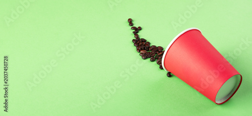 Coffee beans spilling from a red disposable cup on a vibrant green background, perfect for concepts of caffeine, waste, or refreshment. panoramic image