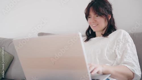 Young woman working on laptop on a cozy sofa in a bright modern living room. Remote work, freelance lifestyle, productivity at home, comfortable workspace, natural light interior.