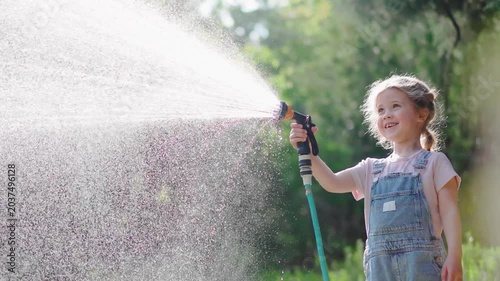 Cheerful little girl watering a green lawn with a garden hose on a warm summer day. Joyful childhood, outdoor play and carefree summer moments in nature.