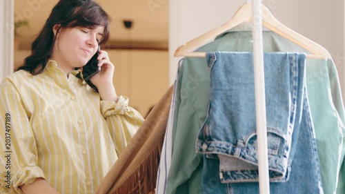 Cheerful young woman in pajamas talking on the phone while choosing an outfit from a clothing rack at home. Morning routine, casual lifestyle and modern female fashion concept.