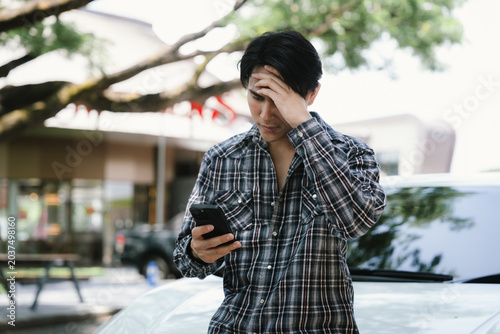Cuadro en lienzo Worried young man looking at a smartphone beside a car, expressing financial stress, unexpected expenses, bad news, problem solving, and personal finance concerns