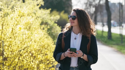 Young student woman with backpack walking through a blooming spring city, checking her phone. Urban student life, campus mobility, youth lifestyle, and modern education concept.